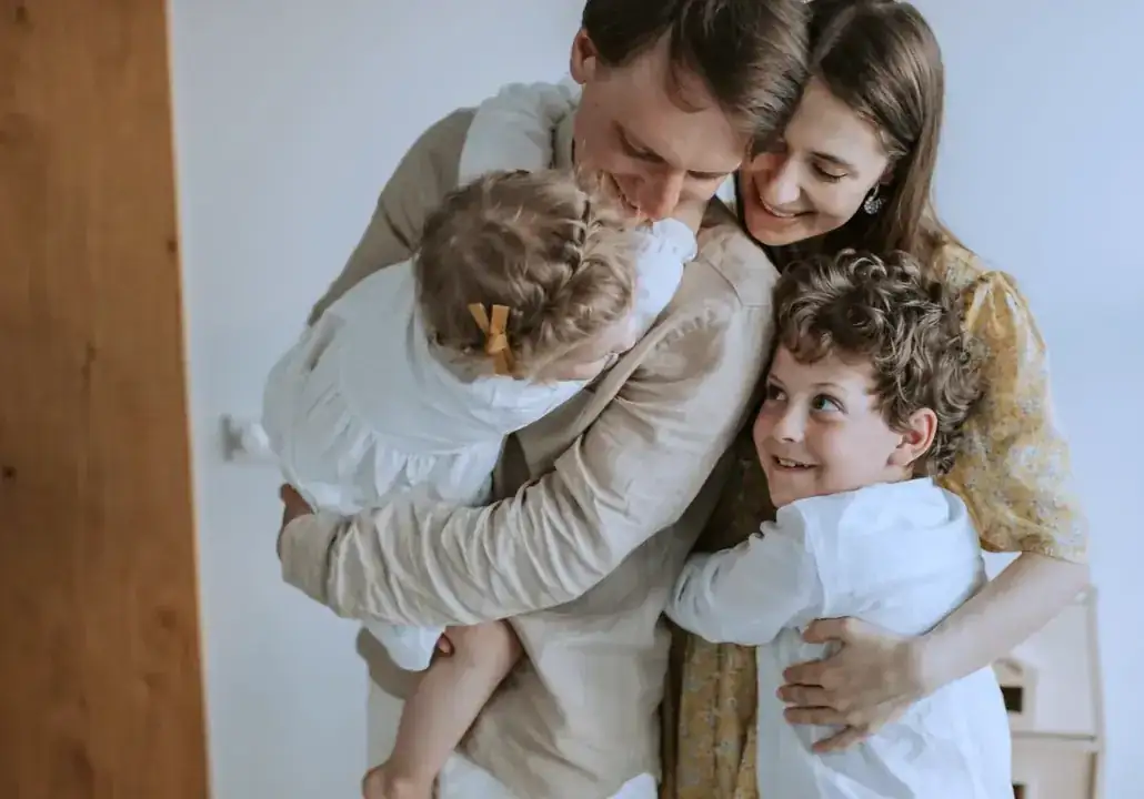 Pai e mãe abraçando os dois filhos pequenos dentro de casa